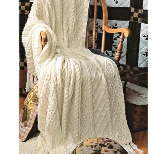 Peut inclure: Plaid de couleur crème en tricot torsadé avec des franges, drapé sur une chaise à bascule en bois. La couverture présente un motif tissé détaillé. Un pouf brodé floral et des coussins décoratifs sont également visibles.