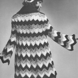 Op de afbeelding: Een zwart, wit en grijs gehaakte jas met chevronpatroon en bijpassende baret. De jas heeft lange mouwen met uitlopende manchetten en een zwarte kraag. De outfit is in vintage stijl.