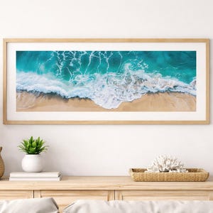 May include: A framed panoramic print of a turquoise ocean with white waves crashing onto a sandy beach. The artwork is in a light wood frame and is displayed above a wooden console table. Decorative items include a lamp, plants, and a woven basket.
