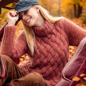 Könnte beinhalten: Ein strukturierter, rostfarbener Pullover mit hohem, geripptem Kragen. Der Pullover hat ein Zopfmuster und lange Ärmel. Eine dunkelblaue Kappe wird getragen. Der Hintergrund zeigt Herbstlaub.