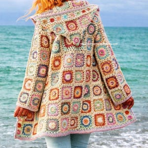 Op de afbeelding: Een lange gehaakte jas met capuchon met een patchwork-ontwerp van kleurrijke vierkanten. Het kleurenpalet van de jas omvat crème, roze, blauw, oranje en groen. De jas wordt gedragen door een persoon die op een strand staat met de oceaan op de achtergrond.