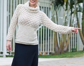 May include: A woman wearing a cream-colored, textured knit sweater with a turtleneck and long sleeves. The sweater is paired with a long, dark navy blue skirt. The woman is smiling and pointing to the right.
