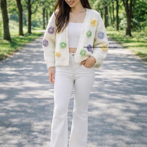 May include: Cream-colored, open-front cardigan adorned with colorful crochet flowers. The sweater features a relaxed fit and is paired with white pants and sneakers. The image includes the Etsy shop URL.
