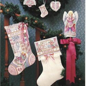 May include: A collection of Christmas stockings and ornaments, featuring cross-stitch designs. The stockings are in pink, white, and blue, with the name "Annabelle" and "Danielle" embroidered on them. Ornaments include a fan, heart, and angel.