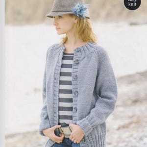 May include: A woman modelling a light blue knitted cardigan with button fastenings, a striped top, and jeans. She wears a grey hat with a blue feather. The image includes the text "CLICK CHUNKY" and "easy knit". The cardigan size is 81-107cm (32-42 inches).