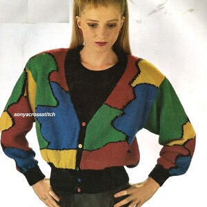 Peut inclure: Un cardigan coloré de style patchwork. Le pull présente un col en V, des manches longues et une fermeture boutonnée. Le design incorpore des formes géométriques rouges, vertes, bleues et jaunes. Les poignets et le col sont noirs. Le texte "sonyacrossstitch" est visible.