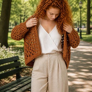 May include: A woman wearing a brown crochet cardigan over a white camisole and beige trousers. The open-weave cardigan has long sleeves and a boxy shape. The trousers are high-waisted with a pleated front. The background is a park with trees and a walking path.