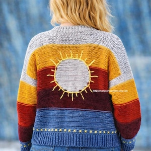 May include: A vibrant, striped jumper with a sun motif. The design includes horizontal stripes in grey, yellow, red, and blue. A yellow sun is embroidered on the back. The sleeves and hem are blue with yellow star details.