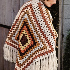 May include: A handmade crochet poncho in a geometric pattern, featuring shades of brown, cream, and orange. The poncho has a fringed edge and is worn over a brown turtleneck. This bohemian style garment is perfect for fall.
