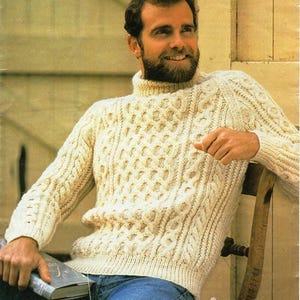 May include: Cream-colored, cable-knit turtleneck sweater. The sweater features a textured pattern with vertical and diamond-shaped designs. The sleeves and body have a ribbed texture. The person is wearing blue jeans.