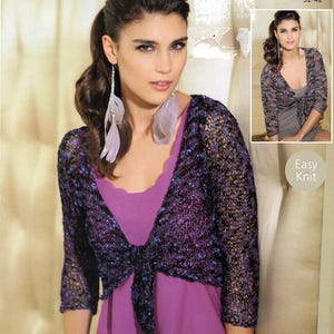 May include: A woman wearing a purple and black knit shrug with a tie closure. The shrug is over a purple top. The image includes the text "FIREFLY" and "9471 81-107cm 32-42". The woman is also wearing feather earrings.