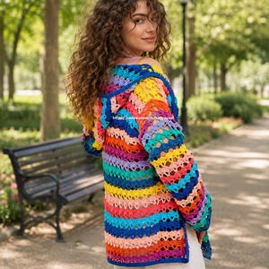 Könnte beinhalten: Ein handgefertigter, gehäkelter Kapuzen-Cardigan in leuchtenden Farben wie Blau, Gelb, Orange und Lila. Das offene Design und die langen Ärmel schaffen ein einzigartiges Kleidungsstück. Die Etsy-Shop-URL ist auf dem Kleidungsstück sichtbar.