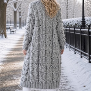 Op de afbeelding: Een lange, grijze, getextureerde gebreide cardigan met een uniek bobbelsteekpatroon. De cardigan is knielang en wordt gedragen over een witte broek en grijze laarzen. De achtergrond toont een besneeuwde buitenomgeving.