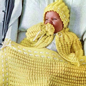 May include: A baby wrapped in a yellow crocheted blanket, wearing a matching hat and cape. The blanket has a textured pattern with white accents along the edges. The hat and cape also feature a textured design.