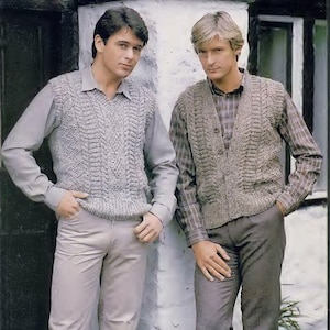 Puede incluir: Dos hombres con chalecos de punto. Uno es gris, el otro marrón. El chaleco gris va sobre una camisa gris con cuello, el marrón sobre una camisa a cuadros. Ambos hombres llevan pantalones.