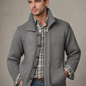 Könnte beinhalten: Grauer Cardigan mit Knopfleiste und Rippstruktur. Der Pullover hat einen Schalkragen, zwei Vordertaschen und wird über einem karierten Hemd und einer blauen Jeans getragen. Ein brauner Gürtel ist sichtbar.