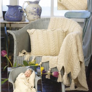 May include: Cream-colored knitted throw blanket and pillow with a cable knit design, tassels, and button details. The blanket is draped over a wicker chair. A small inset image shows a child wrapped in a matching blanket.