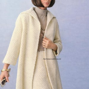 May include: A vintage cream-colored textured coat and skirt ensemble. The coat has a long, flowing design with a wide collar. The outfit is paired with a turtleneck and a gold necklace. The model is also wearing a gold bracelet and holding sunglasses.