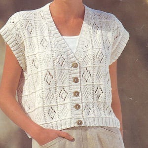 May include: Cream-colored, short-sleeved cardigan with a V-neck and button closure. The knitted garment features a geometric diamond pattern and ribbed edges. The buttons are a light brown color. The cardigan is worn over a white tank top.