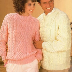May include: A couple in matching cable-knit sweaters. The woman's sweater is pale pink, and the man's is cream. Both sweaters have a high neckline and a textured pattern. The woman is wearing pale pink trousers.