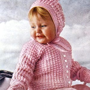 May include: A baby wearing a pink crochet outfit, including a bonnet with a bow, a button-up sweater, and a matching scarf. The outfit has a textured pattern and is made of a soft, possibly wool, material. The baby is smiling.