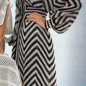 May include: A long, open-front dress with a chevron pattern in black and white. The dress has long sleeves and a V-neckline. The model is wearing a white top underneath. The dress is made of a knitted material.