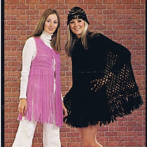May include: Two women modeling handmade crochet clothing. One wears a long, pink fringed vest over a white turtleneck and pants. The other wears a black crochet poncho with a matching hat. The background is a brick wall.
