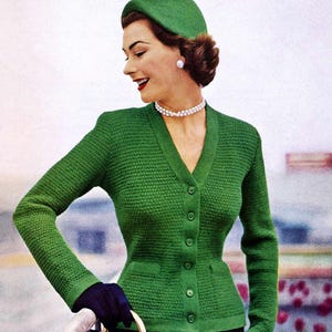 May include: A woman in a green knitted cardigan, hat, and pearl necklace. The cardigan features a V-neck, button fastening, and small pockets. She holds a navy handbag with a gold handle. The woman is also wearing black gloves.