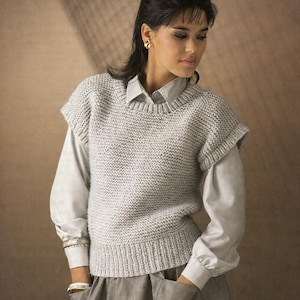 May include: A light gray knitted vest with a ribbed hem and short sleeves. The vest is worn over a light gray collared shirt and a gray skirt. The model is wearing gold earrings and a bracelet.