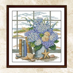 May include: A framed cross-stitch embroidery depicting a window view of a beach scene with a bouquet of blue hydrangeas and yellow roses in a metal bucket, surrounded by seashells and a starfish.