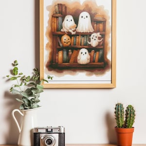 May include: A framed print featuring a bookshelf filled with books and several cartoon ghosts. A small pumpkin sits on the shelf. The print is displayed on a wooden surface with a vintage camera, a white pitcher with greenery, and a potted cactus.