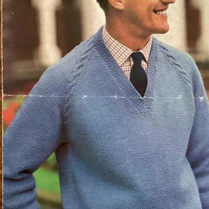 以下が含まれることがあります： 肩にケーブルニットのディテールが施された、ライトブルーのVネックセーター。セーターは襟付きシャツと濃い青色のネクタイの上に着用されています。画像には笑顔の男性が写っています。