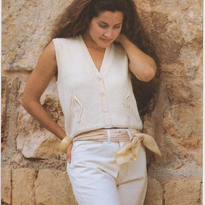 May include: Cream-coloured knitted vest with a V-neck and button closure. The vest has decorative openwork details on the front. Worn with off-white jeans and a matching scarf tied around the waist. A neutral-toned outfit.