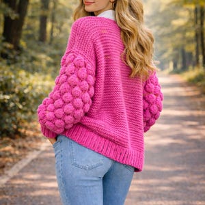 May include: A bright pink knit sweater with bubble sleeves. The sweater is paired with light blue jeans. The model is outdoors on a path surrounded by trees.
