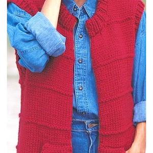 May include: A red knitted vest with a full-length zipper and two front pockets. The vest is worn over a denim shirt and blue jeans. The knitted texture is visible, and the vest appears to be handmade.