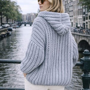 Puede incluir: Un suéter con capucha de punto grueso gris claro con una textura acanalada. El suéter tiene un ajuste relajado y se combina con pantalones blancos. La persona lleva gafas de sol y está de pie cerca de un canal con edificios al fondo.