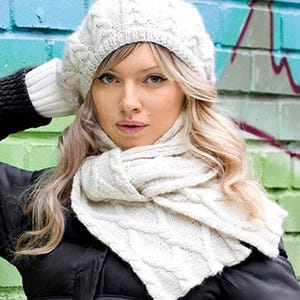 May include: A woman wearing a white cable-knit beret, scarf, and gloves. The winter accessories are made of a soft, textured yarn. The woman is wearing a black jacket. The background is a painted brick wall with graffiti.