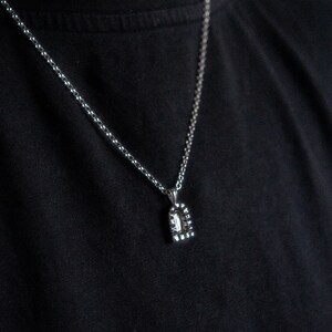 May include: A silver chain necklace with a small, silver pendant. The pendant is shaped like a rectangle with rounded corners and has a textured surface.