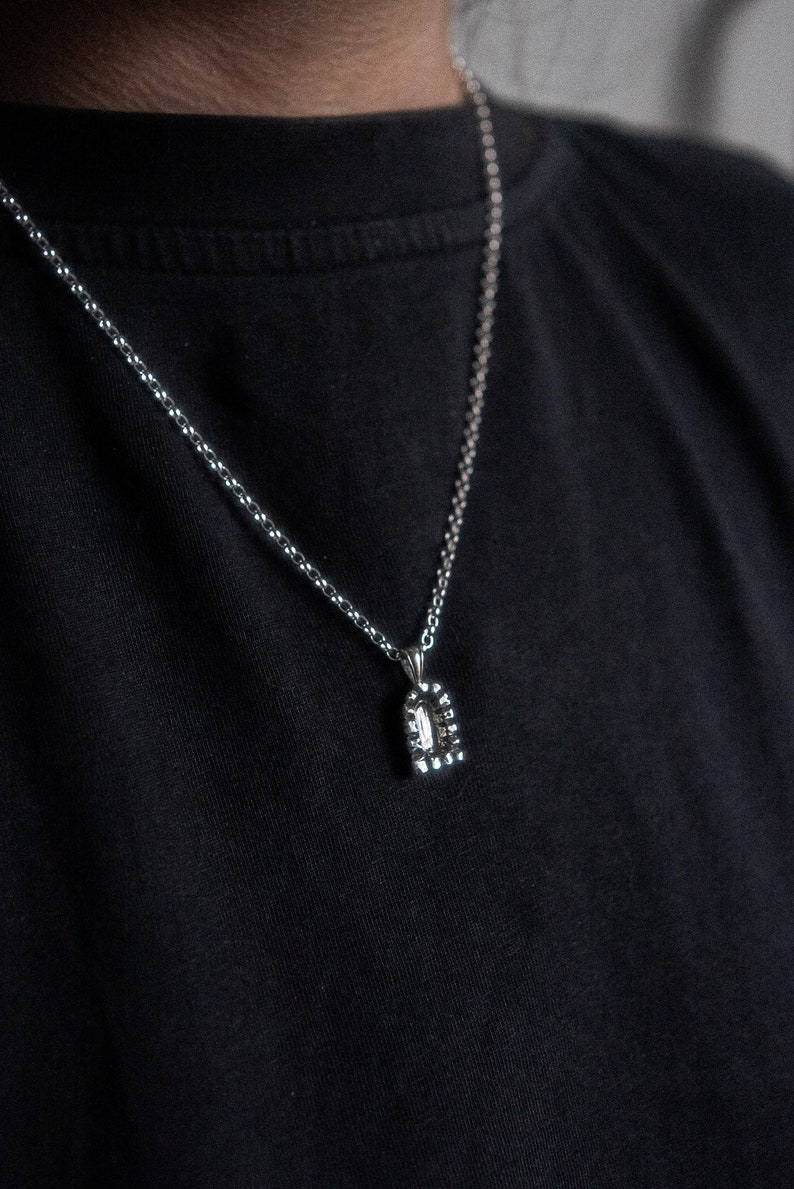 May include: A silver chain necklace with a small, silver pendant. The pendant is shaped like a rectangle with rounded corners and has a textured surface.