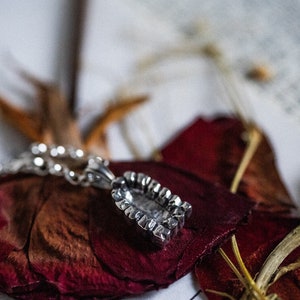 May include: A silver pendant necklace with a detailed, textured design. The pendant is shaped like a small, rectangular box with a raised edge. The necklace is lying on top of dried rose petals.