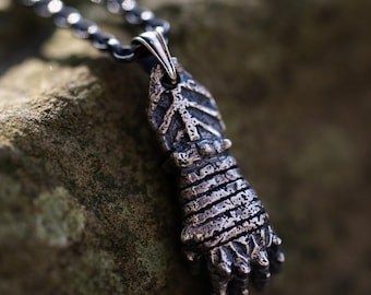 Puede incluir: Un collar de plata con un colgante texturizado detallado en forma de mano con garras. El colgante cuelga de una cadena sobre un fondo marrón texturizado.