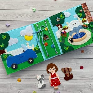 Puede incluir: Un libro de fieltro con un coche azul sobre un paisaje verde, un panel central verde con cordones azules y una escena de jardín con fuente. Las figuras de fieltro sueltas incluyen una persona con vestido rojo, un gato gris y blanco y un perro marrón.