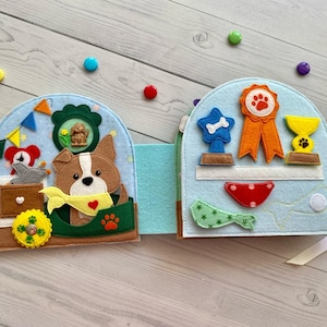 May include: A light blue felt quiet book featuring a brown and white dog with a yellow bandana on the left page, and a blue star trophy, orange ribbon, and yellow trophy on the right page, decorated with colorful felt shapes and small buttons.