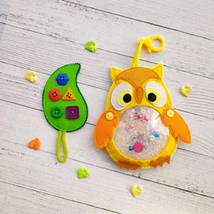 May include: A yellow felt owl sensory pouch with a clear plastic belly filled with colorful beads and a green felt leaf-shaped accessory featuring various plastic buttons in blue, red, orange, and purple, accompanied by small green felt leaf accents.