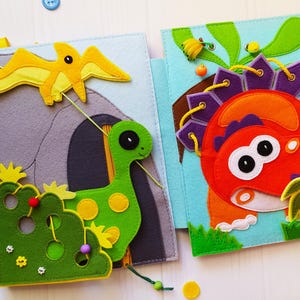 May include: A felt quiet book featuring dinosaur-themed pages. One page shows a yellow pterodactyl, a green long-necked dinosaur, and a gray cave. Another page displays a red and orange dinosaur with purple spikes and yellow lacing details.
