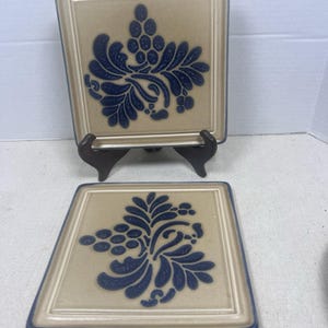 May include: Two square ceramic tiles featuring a tan base color with a dark blue embossed grape and leaf pattern. The tiles have a raised border edge and a smooth finish. One tile is displayed upright on a black stand, while the other lies flat.