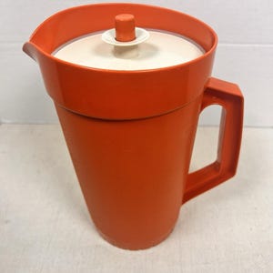 May include: An orange plastic pitcher with a white lid and handle. The pitcher has a textured surface and a spout for pouring. The lid has a small orange handle. The pitcher is likely vintage and suitable for serving beverages.