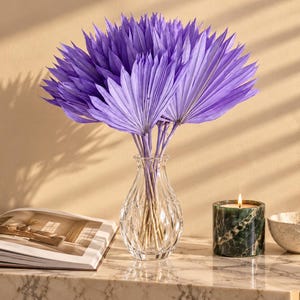 Könnte beinhalten: Ein Strauß aus violetten, getrockneten Palmblättern in einer klaren, gerippten Glasvase. Neben der Vase stehen ein dunkelgrüner Marmorkerzenhalter mit brennender Kerze und eine kleine, strukturierte cremeweiße Keramikschale neben einem Buch.