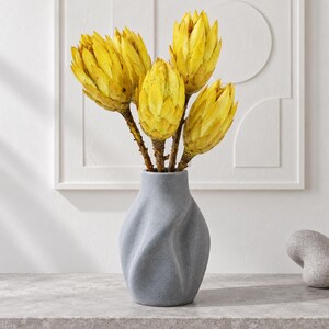 May include: A gray ceramic vase with a twisted, textured surface containing four bright yellow dried protea flowers. The vase has a narrow neck and a wider, rounded base, holding the stems of the large, pointed, golden-yellow floral blooms.