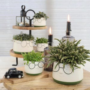 Könnte beinhalten: Eine Anordnung von weißen Keramik-Pflanztöpfen mit grünen Böden, jeweils mit einem lächelnden Gesicht und runden Brillen verziert. Die Töpfe enthalten verschiedene grüne Pflanzen und sind mit Kerzen und einem kleinen schwarzen Spielzeugauto auf einer Holzoberfläche angeordnet.